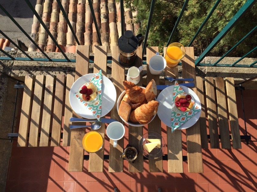A breakfast table laid on the terrace at Villa Rocamar: fruit, yoghurt, granola and juice, croissant and baguette with coffee.